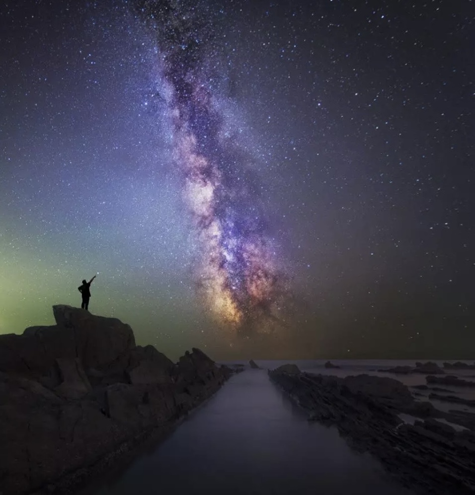 A person standing on a rock at night, arm raised toward the Milky Way above a dark shoreline.